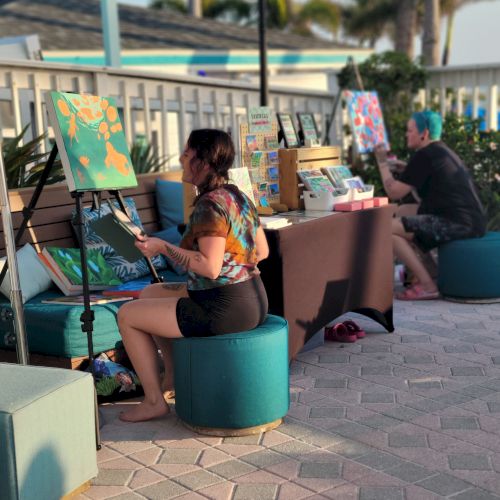 Two people are painting on canvases outdoors. One person is seated in the foreground while another is painting in the background, with palm trees around.