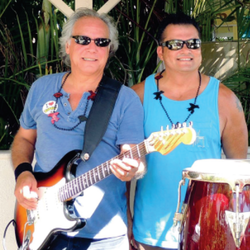 Two men are smiling and standing together outdoors; one is holding an electric guitar, and the other is standing next to congas.