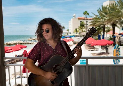 A person with long hair and sunglasses is playing a guitar on a beachside patio, with red umbrellas and palm trees in the background.