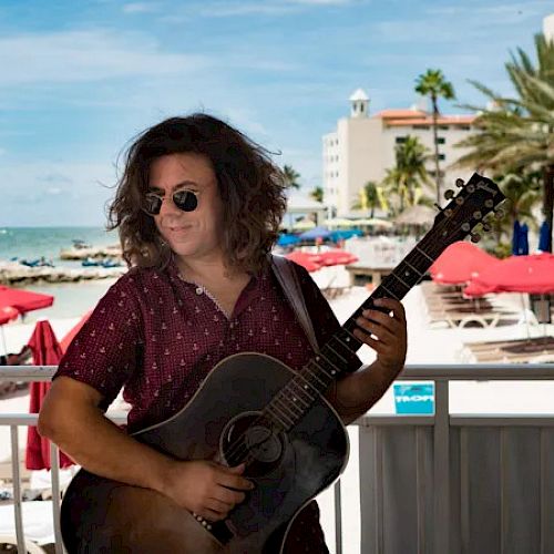 A person with long hair and sunglasses is playing a guitar on a beachside patio, with red umbrellas and palm trees in the background.