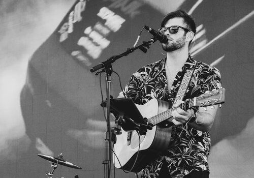 A person wearing sunglasses and a patterned shirt is playing guitar and singing into a microphone on stage with a drum set in the background.