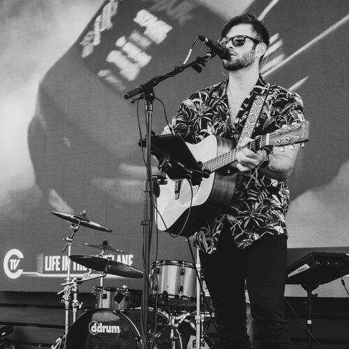 A person wearing sunglasses and a patterned shirt is playing guitar and singing into a microphone on stage with a drum set in the background.