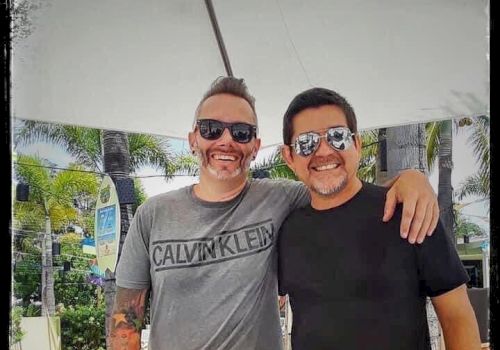 Two people are smiling and posing together outdoors under a shade. They are both wearing sunglasses and casual clothing.