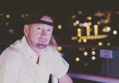 A man in a white shirt and cap sits on a railing with a nighttime cityscape in the background, featuring blurred lights.