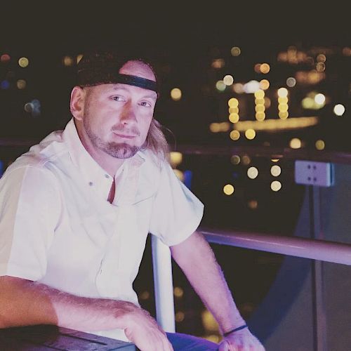 A man in a white shirt and cap sits on a railing with a nighttime cityscape in the background, featuring blurred lights.