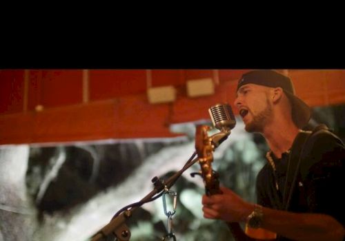 A man wearing a backwards cap is singing into a vintage microphone while playing an acoustic guitar in a dimly lit room.