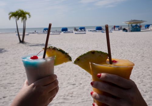 Two hands holding tropical drinks with pineapple garnishes on a sunny beach with umbrellas and lounge chairs in the background.