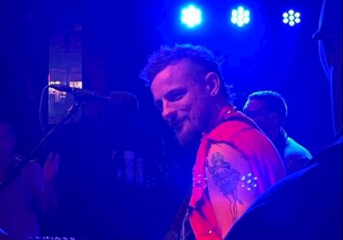 A musician wearing a red vest playing guitar on stage, illuminated by blue stage lights, with other musicians and audience members in the background.