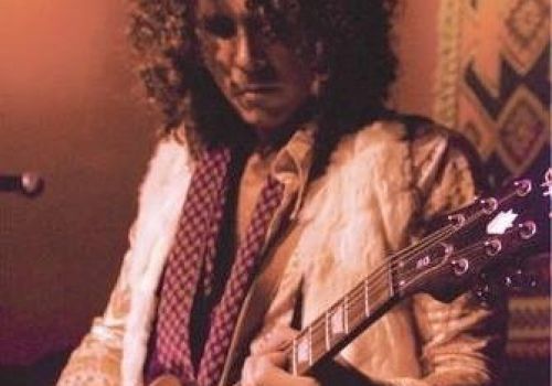 A person with curly hair plays an electric guitar, wearing a patterned jacket and scarf, against a warm, dimly lit background with decorative elements.