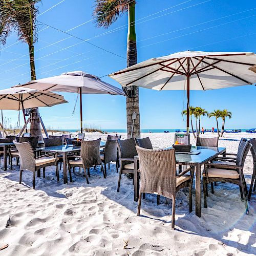 Outdoor beachside dining area with tables, chairs, and umbrellas on white sand, with the ocean and palm trees in the background.