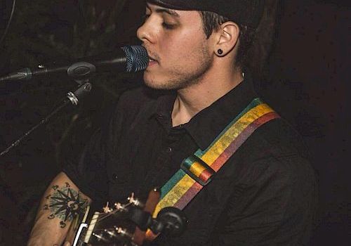 A person wearing a hat and black shirt is playing a guitar and singing into a microphone, with a colorful guitar strap around their shoulder.