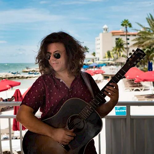 A person with long hair, wearing sunglasses and a red shirt, is playing a guitar on a beach with palm trees and red umbrellas in the background.