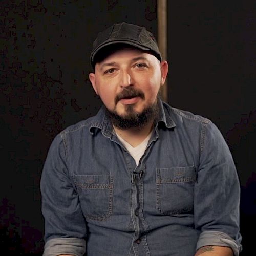 A person wearing a denim shirt and a cap is sitting against a dark background, looking towards the camera.