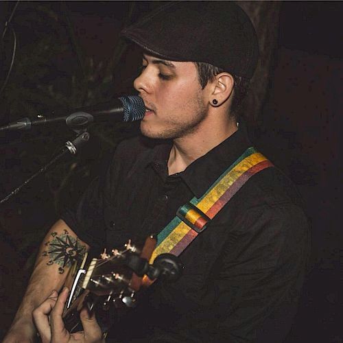 A person wearing a dark outfit and cap is singing into a microphone while playing a guitar with a colorful strap.