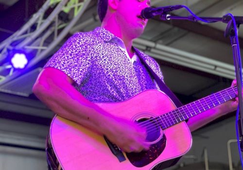 A musician plays an acoustic guitar and sings into a microphone on a stage with colorful lights overhead.
