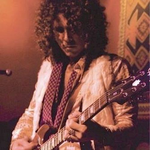 A person with curly hair plays an electric guitar, wearing a patterned jacket and scarf, against a warm, dimly lit background with decorative elements.