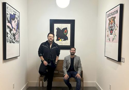 Two men in a narrow room, one standing and one sitting on a bench, with artwork on the walls and a hanging light above them.